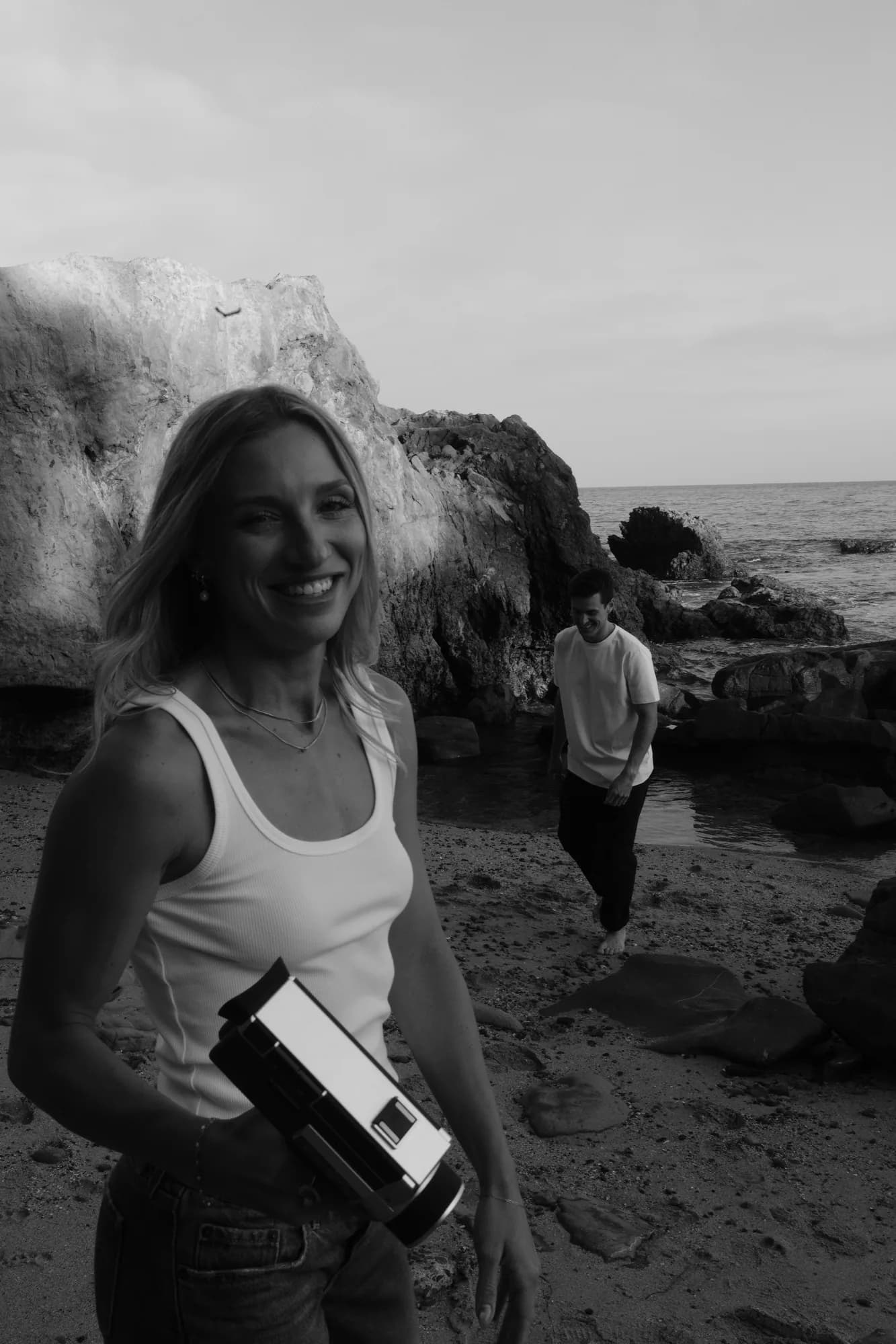 Smiling woman holding a vintage film camera on the beach with her partner behind her, black and white