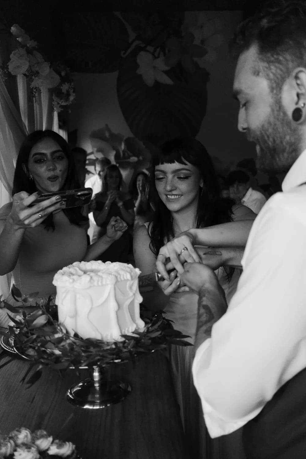 Couple cutting their wedding cake with friends looking on, black and white