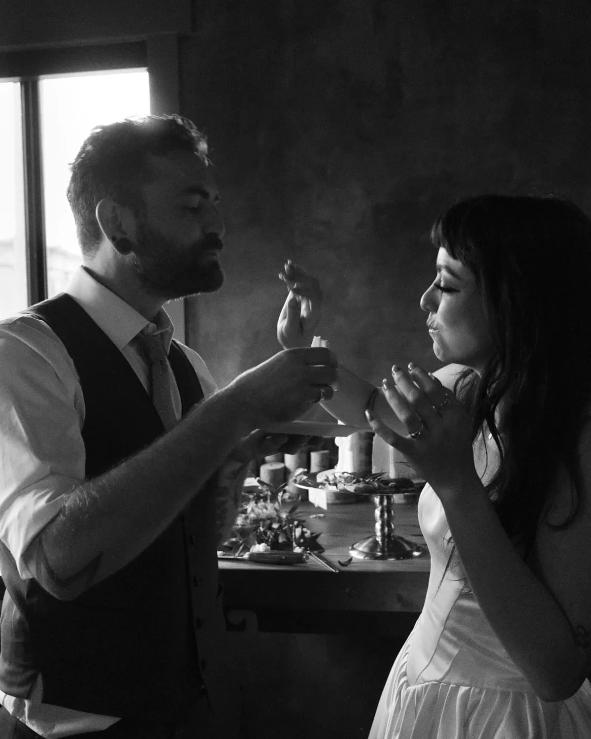 Couple sharing cake in soft window light at their intimate wedding, black and white