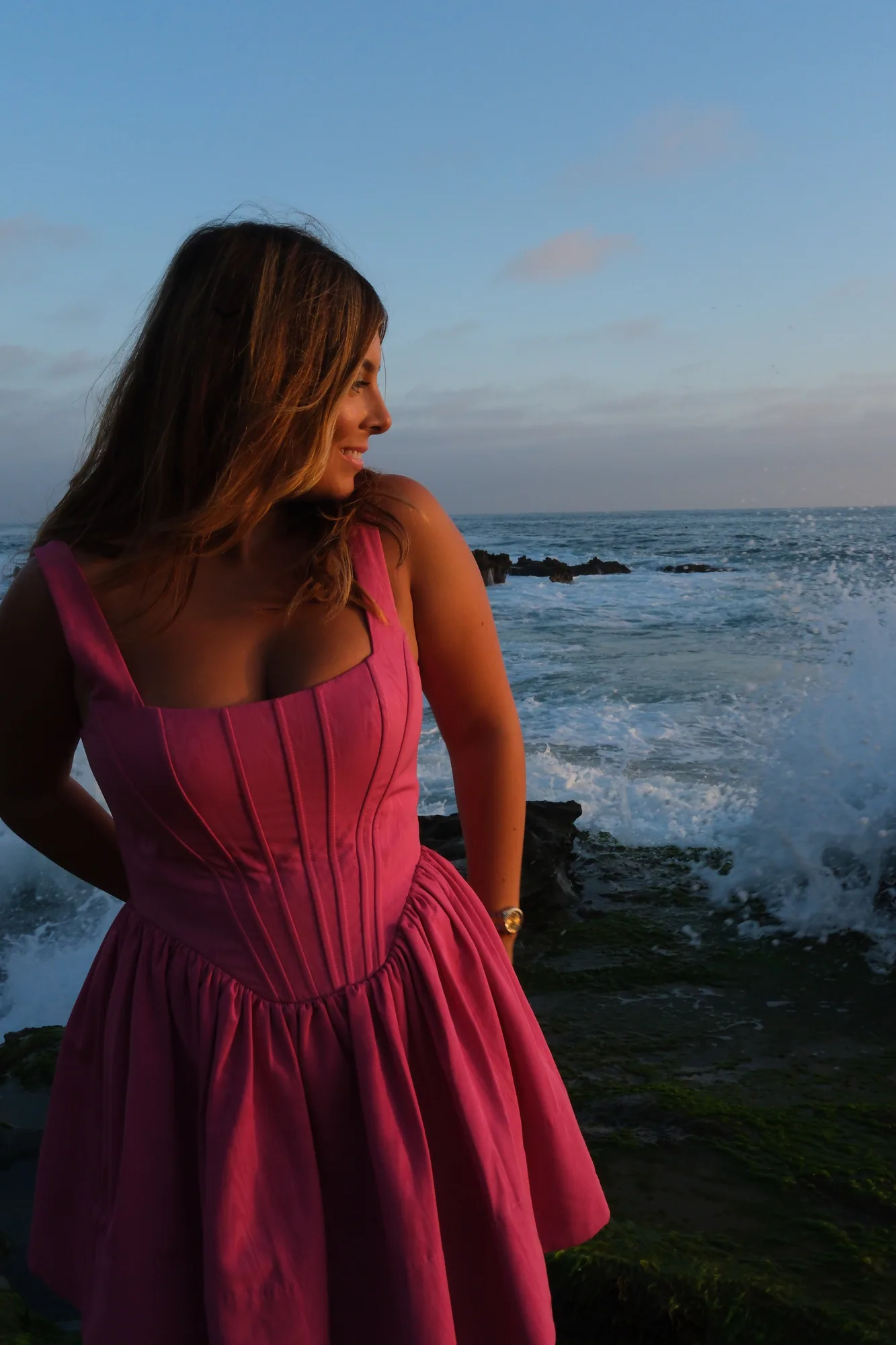 Young woman in a pink dress looking out at ocean waves during a golden hour portrait session