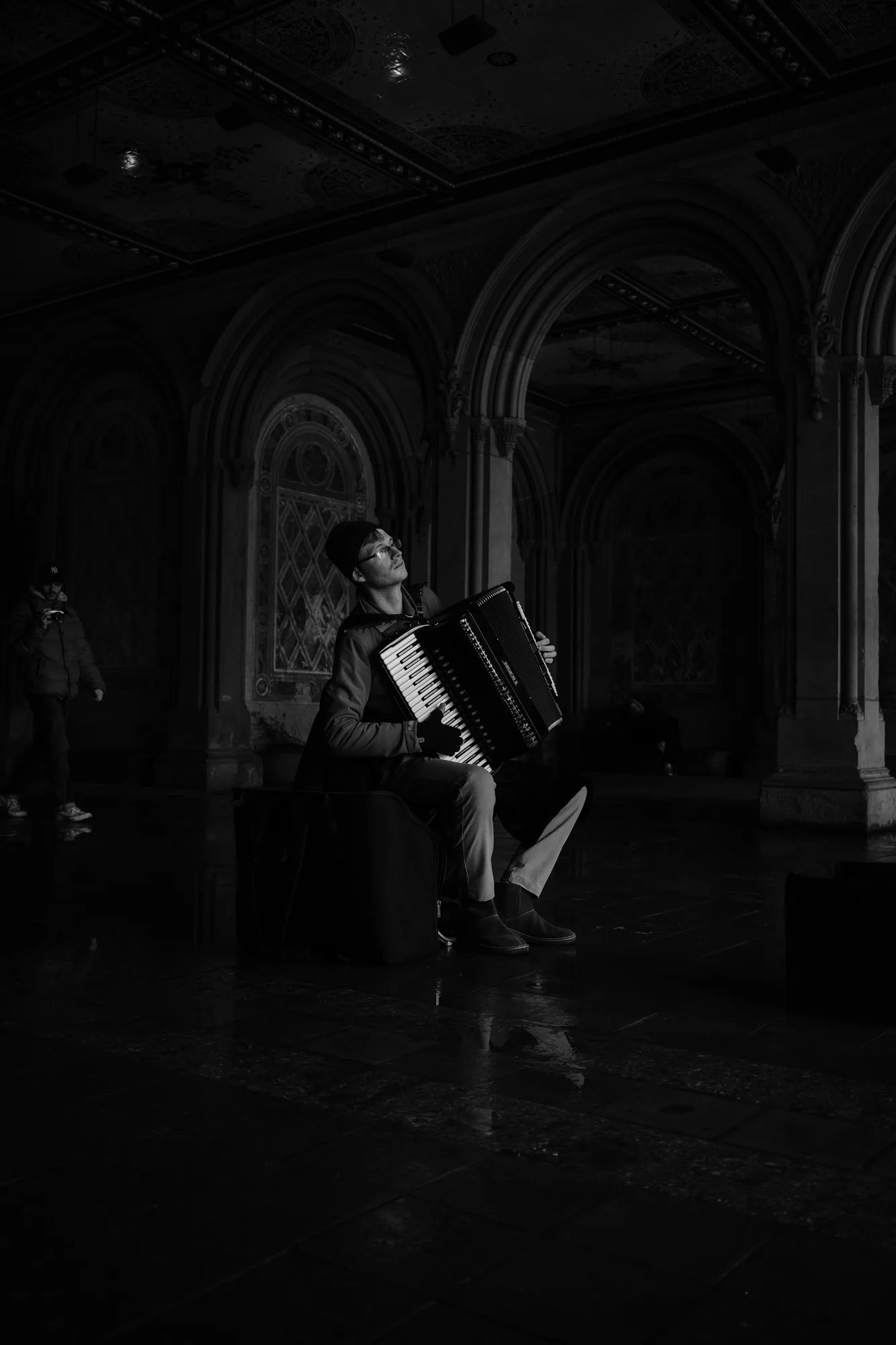 Black and white photo of a musician playing an accordion in a vaulted arcade in New York City