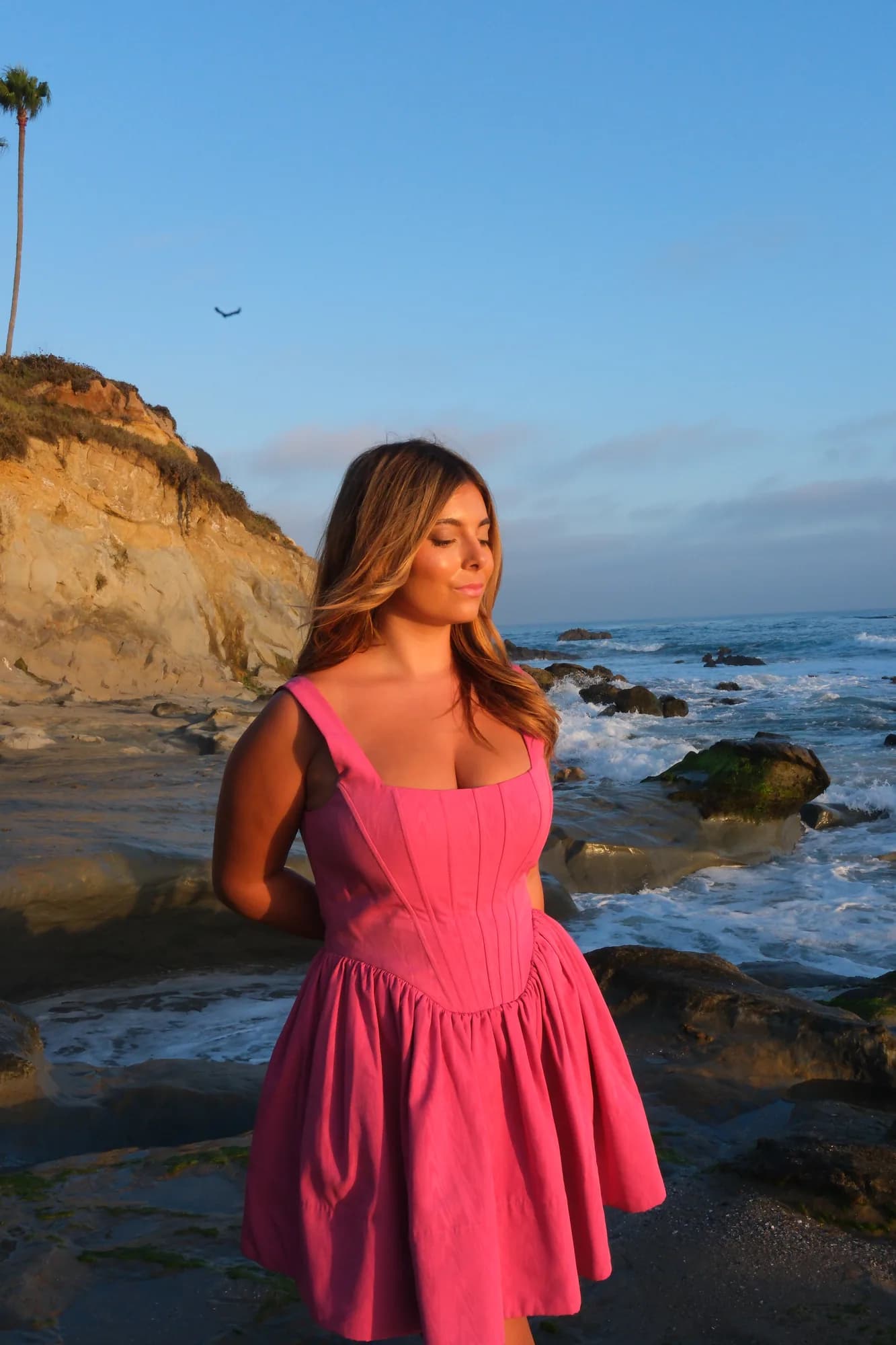 Young woman in a pink dress standing on rocks below coastal cliffs at sunset